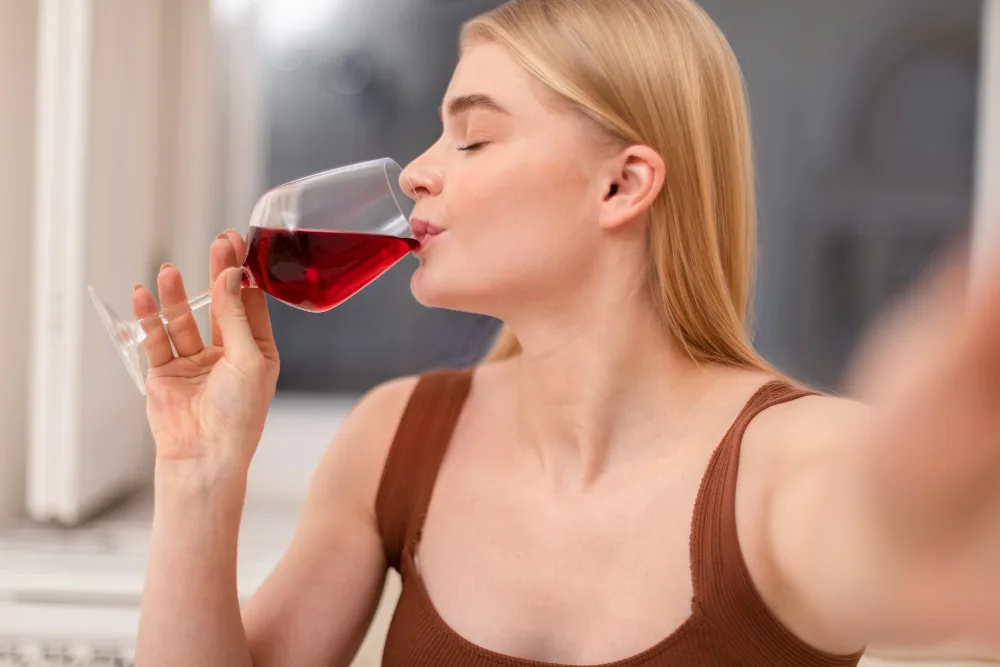 Woman with blonde hair in a brown sleeveless top drinks red wine from a stemmed glass, eyes closed in a bright indoor setting.