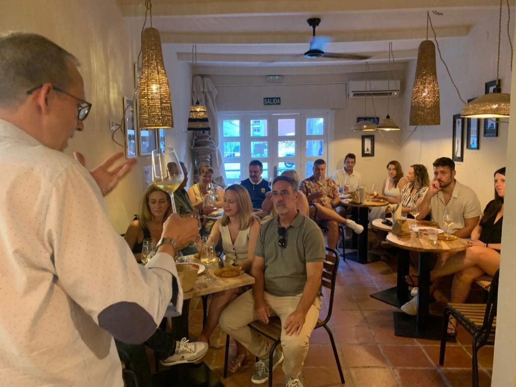 A host pours a glass of wine for a group dining in a cozy restaurant, guests seated at round tables with bread and glasses nearby.