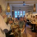 A host pours a glass of wine for a group dining in a cozy restaurant, guests seated at round tables with bread and glasses nearby.