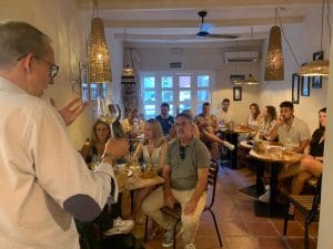 A host pours a glass of wine for a group dining in a cozy restaurant, guests seated at round tables with bread and glasses nearby.