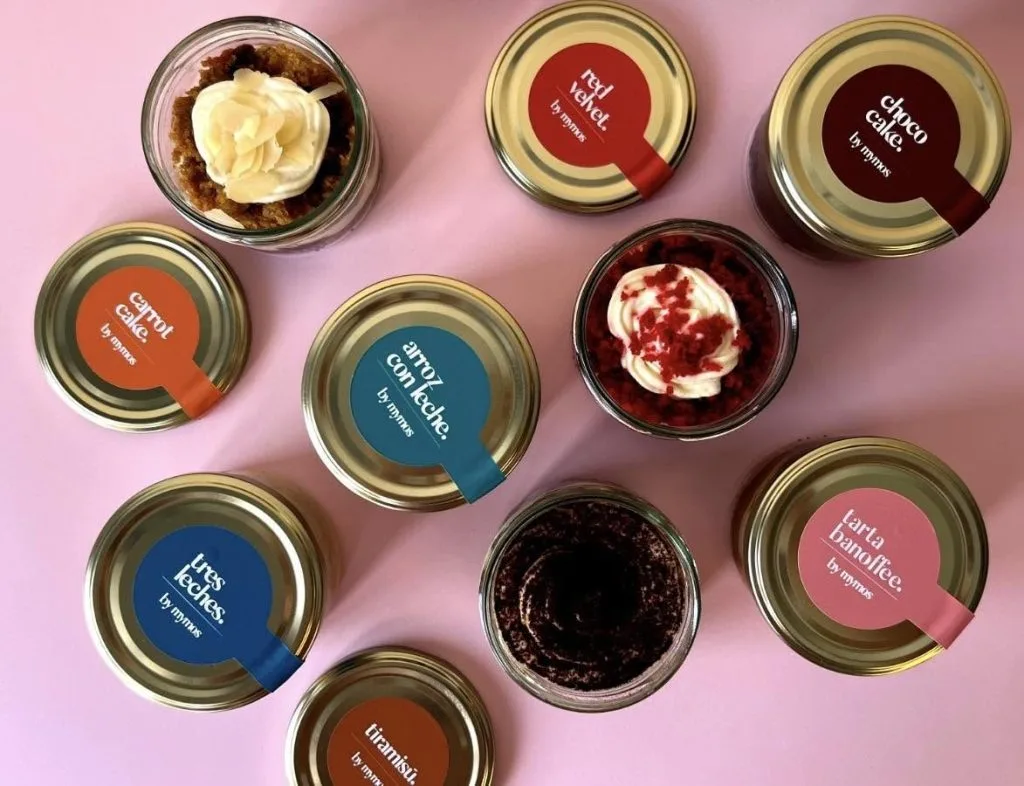 Top-down view of multiple dessert jars with color-coded labels arranged on a pink background, showing assorted flavors in glass jars with metal lids.