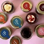 Top-down view of multiple dessert jars with color-coded labels arranged on a pink background, showing assorted flavors in glass jars with metal lids.