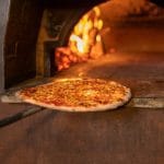 Pizza on a long paddle sliding toward a wood-fired oven with glowing flames in the background, ready to bake.