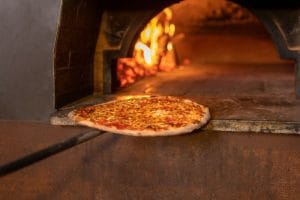 Pizza on a long paddle sliding toward a wood-fired oven with glowing flames in the background, ready to bake.