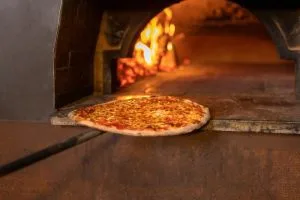 Pizza on a long paddle sliding toward a wood-fired oven with glowing flames in the background, ready to bake.