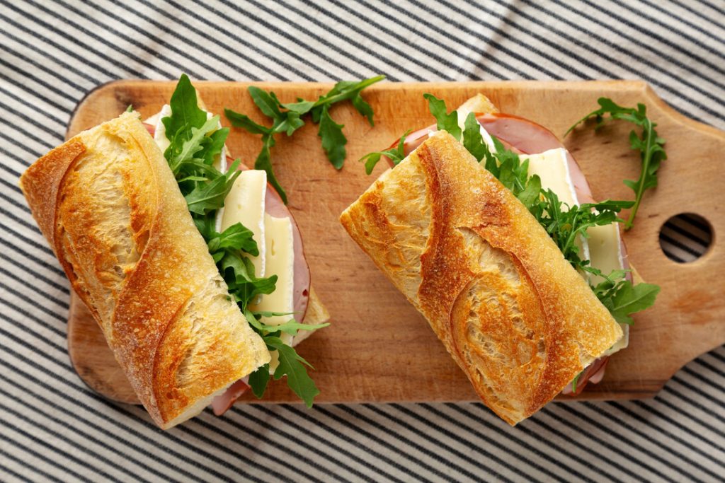 Homemade French Ham and Brie Baguette with Arugula Greens top view - Recetitas.Com Two baguette sandwiches filled with arugula, cheese and ham on a wooden cutting board, sliced diagonally open for serving
