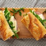 Two baguette sandwiches filled with arugula, cheese and ham on a wooden cutting board, sliced diagonally open for serving